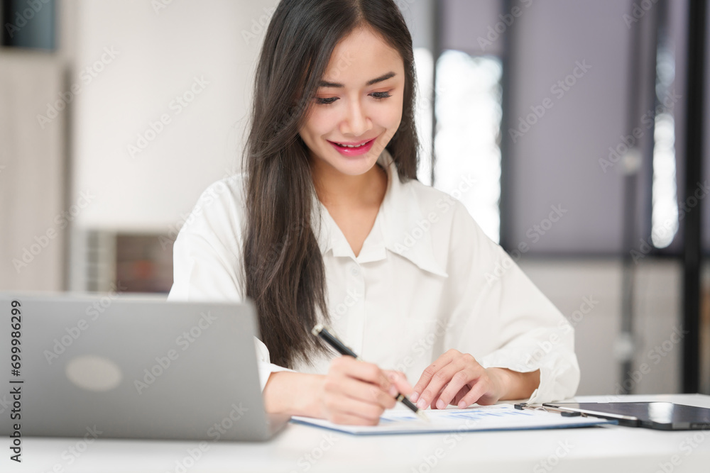 young Asian business woman with long hair writing on paper at a desk with a laptop open in front of her. young Asian business woman with long hair writing on paper at a desk with a laptop open in front of her.
