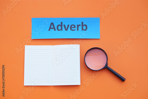 Paper word card with text Adverb, magnifying glass and paper notebook. Orange background. Concept, English grammar teaching. Learning English language lesson. 
