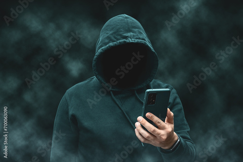 Mysterious faceless hooded anonymous hacker holding smartphone, silhouette of cybercriminal, terrorist