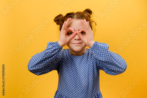 Curious child exploring world. Portrait of inquisitive nosy little girl kid looking through fingers shaped like binoculars and expressing amazement, posing isolated over yellow studio background wall