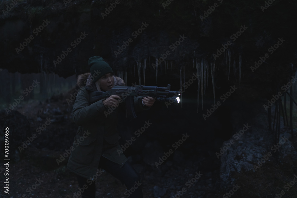Fototapeta premium A civilian girl with a machine gun and a flashlight takes aim at night in the dark in the forest near the ruins, with weak lighting.
