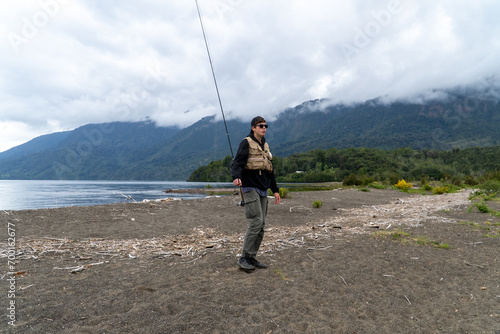 Young Hispanic man walking with a fishing rod by the lake on a cloudy day.