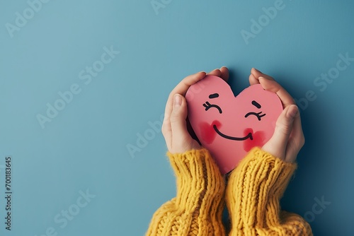 Female hands hold a paper heart with a smiley face isolated over blue background. world heart day, health care, love, organ donation, world health day, wellbeing, gratitude, be kind, be thankful, pray