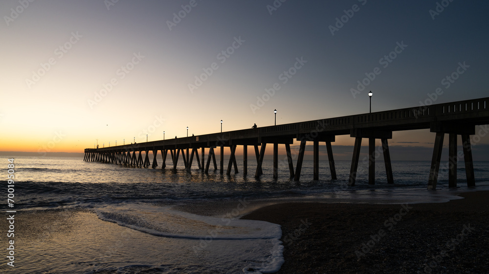 Sunrise illuminates a pier at Wrightsville Beach, casting long shadows over the ocean waves. Sunrise illuminates a pier at Wrightsville Beach, casting long shadows over the ocean waves.