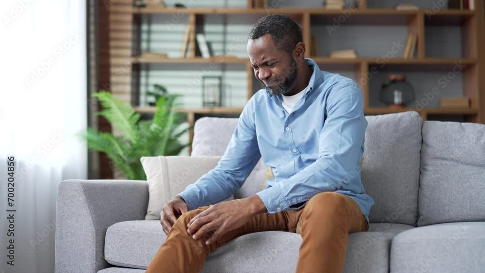 Mature african american man suffers from painful severe knee joint pain sitting on sofa in living room at home. Upset senior male massages leg muscles, could not walk, illness, cramps, rheumatism Mature african american man suffers from painful severe knee joint pain sitting on sofa in living room at home. Upset senior male massages leg muscles, could not walk, illness, cramps, rheumatism