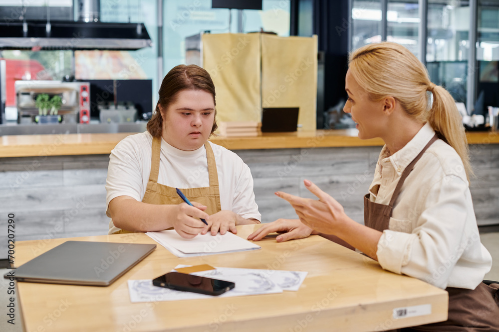 Fototapeta premium cafe administrator talking to female employee with down syndrome writing near laptop and smartphone