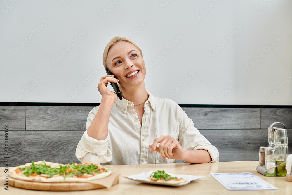 happy blonde woman messaging on smartphone near delicious pizza on table in modern cozy cafe