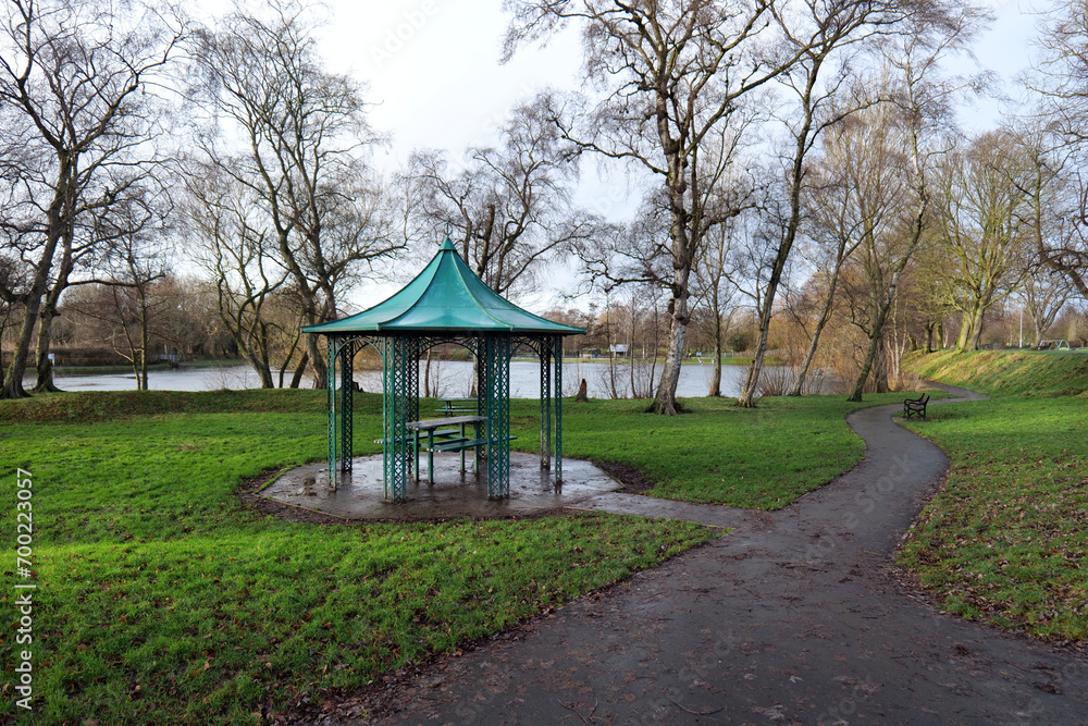 A green metal shelter covers a picnic seating area in a public park. Trees and grass are around it and a boating lake can be seen in the background. Shot in winter with a grey sky