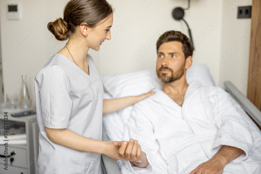 Young nurse takes care of adult man lying on bed of comfortable medical ward. Medical support and assistance during the rehabilitation in clinic