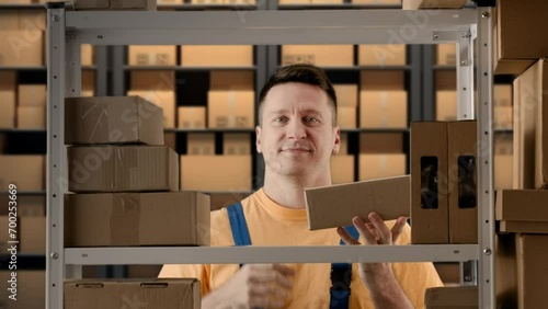 Portrait of male working in storage. Man storekeeper standing near rack with boxes, holding package in hand shows thumbs up at camera. photo