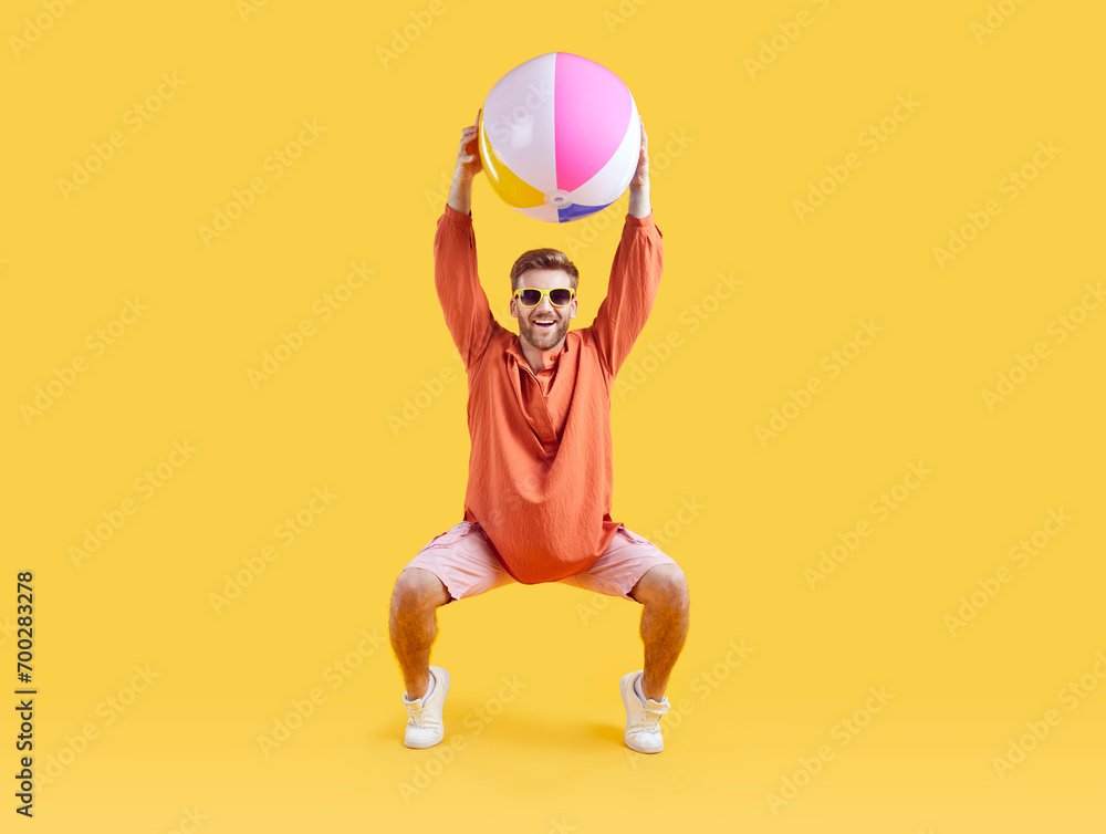 Happy young man exercising with inflatable sea ball. Full length shot of excited man wearing summer casual clothes and sunglasses having fun over isolated yellow studio background Happy young man exercising with inflatable sea ball. Full length shot of excited man wearing summer casual clothes and sunglasses having fun over isolated yellow studio background