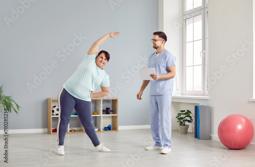 Positive overweight young woman doing sports exercises under supervision of medical worker. Friendly male physiotherapist is helping fat woman in rehabilitation center. Concept of medicine.