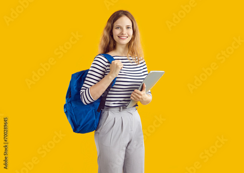 Studio shot of happy student girl who studies at college or university. Smiling young woman in casual top and trousers, with blue backpack and notebook standing isolated on yellow colour background