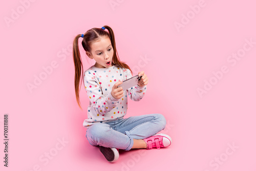 Full size photo of impressed schoolgirl dressed dotted pullover sit play in video game on smartphone isolated on pink color background