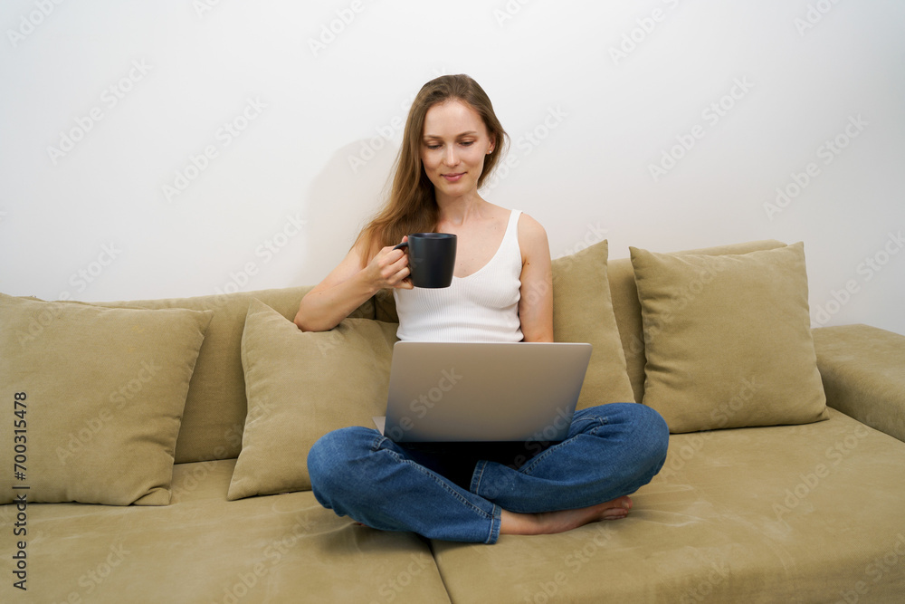 A young woman drinks tea from a cup and works on a laptop while sitting on a sofa. The concept of remote work, watching movies and online courses. A young woman drinks tea from a cup and works on a laptop while sitting on a sofa. The concept of remote work, watching movies and online courses.