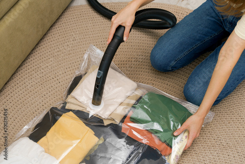 A woman uses a vacuum cleaner to extract air from a transparent vacuum bag with clothes while sitting on the floor. The concept of compact storage and saving space in the closet
