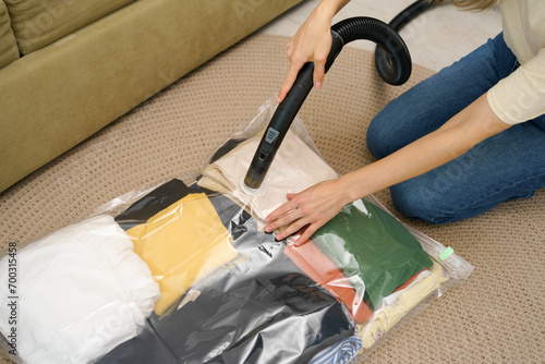 A young woman uses a vacuum cleaner to extract air from a vacuum bag with clothes for compact storage in a closet while sitting on the floor. Space saving concept, compressed packaging and seal bag photo