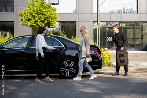 Female chauffeur opens a car door for a business lady going to sit, man packs a suitcase into a car trunk. Concept of luxury car transfer service, business trips and taxi