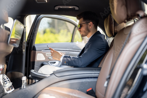 Businessman sits with phone on a backseat of luxury car. Man doing business on the road in prestigious car. Concept of business transportation