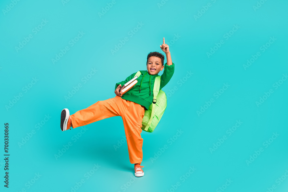 Full size photo of small boy with rucksack wear green hoodie hold book directing up at empty space isolated on turquoise color background Full size photo of small boy with rucksack wear green hoodie hold book directing up at empty space isolated on turquoise color background