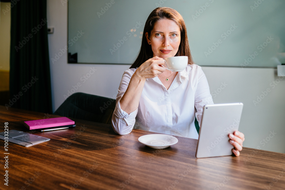 Smiling businesswoman using tablet computer holding cup of coffee looking at camera. A mature female employee working on a report or project at desk in office. Smiling businesswoman using tablet computer holding cup of coffee looking at camera. A mature female employee working on a report or project at desk in office.