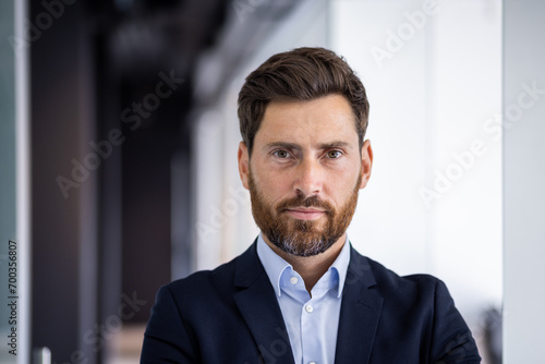 Close-up portrait of a serious young successful male businessman standing in the office in a suit and confidently looking at the camera