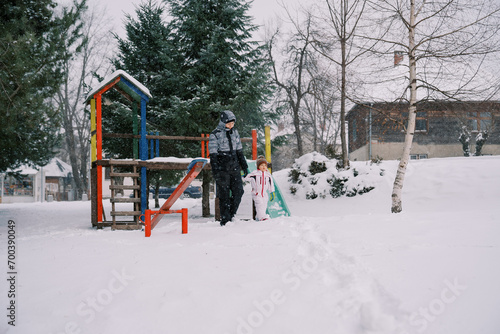 Little girl and her mother walk holding hands from a colorful snow-covered slide through the park under snowfall