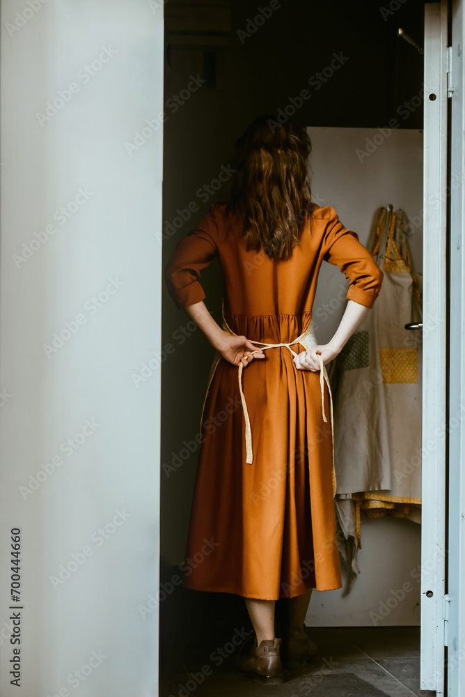 Woman standing at the front door tying an apron. Concept of starting a new day, unplugging, creativity. Master preparing for a new day in the workshop