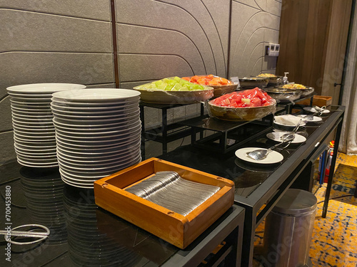 Buffet table setting in hotel or restaurant. Stack of plates, pile of spoons, wrapped cut fruits, grey textured wall photo