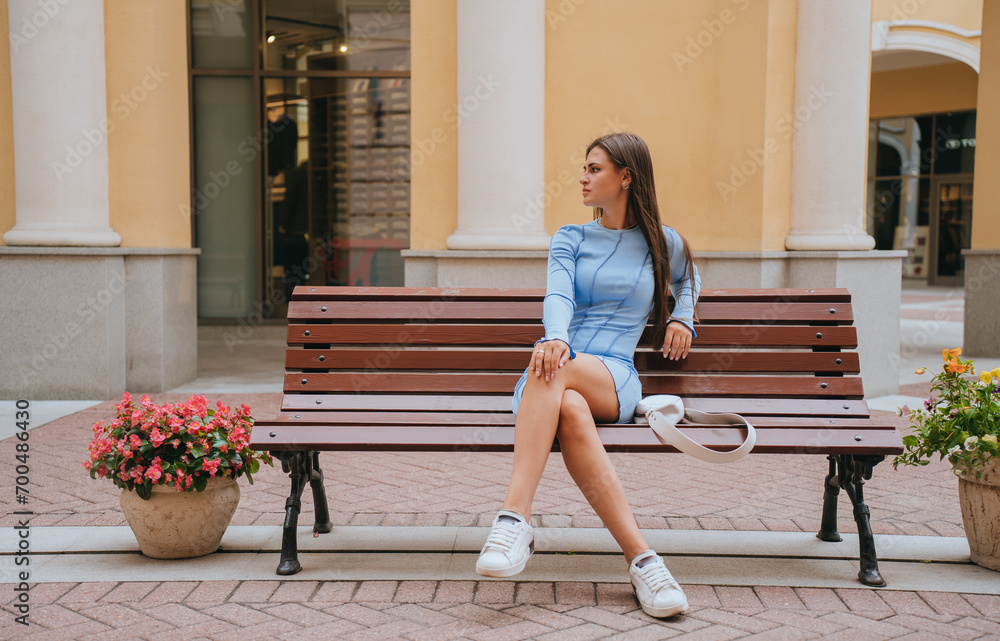 Chic woman in a blue dress and white sneakers sits on a city bench, with a stylish bag beside her and colorful flowers nearby