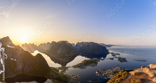 The midnight sun casts its ethereal glow over a super panorama seen from Reinebringen; a wide-angle perspective shows the dramatic Lofoten peaks surrounding the mirror-like fjord with Reine village
