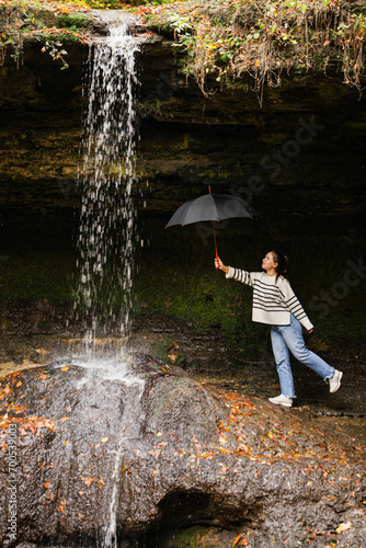 Happy young girl holding black umbrella under the drops of flowing waterfall, looks up to the waterfall and smiles. concept of visiting the most beautiful waterfalls.