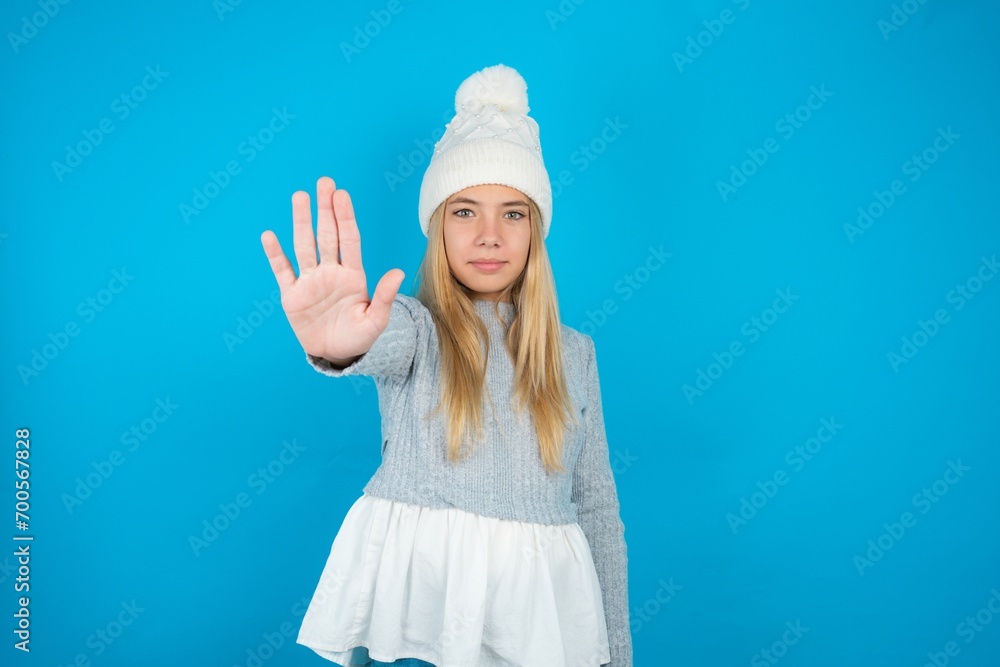 Caucasian blonde girl wearing blue sweater and wool beanie doing stop sing with palm of the hand. Warning expression with negative and serious gesture on the face. Caucasian blonde girl wearing blue sweater and wool beanie doing stop sing with palm of the hand. Warning expression with negative and serious gesture on the face.
