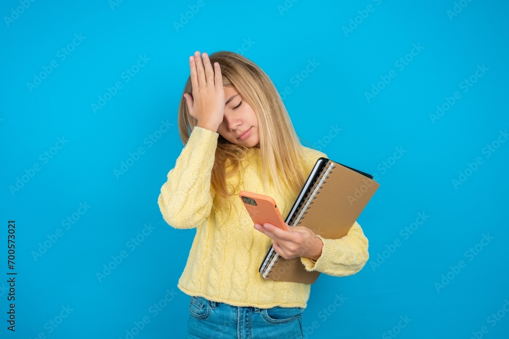 Upset depressed beautiful caucasian teen girl wearing yellow sweater makes face palm as forgot about something important holds mobile phone expresses sorrow and regret blames Upset depressed beautiful caucasian teen girl wearing yellow sweater makes face palm as forgot about something important holds mobile phone expresses sorrow and regret blames