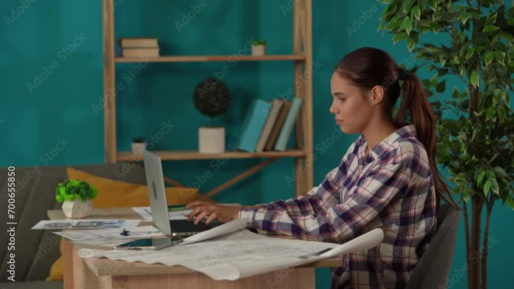 Medium shot capturing a woman working in her home office. Young architect looking through, comparing the blueprints of the building and inserting data into her laptop. Medium shot capturing a woman working in her home office. Young architect looking through, comparing the blueprints of the building and inserting data into her laptop.