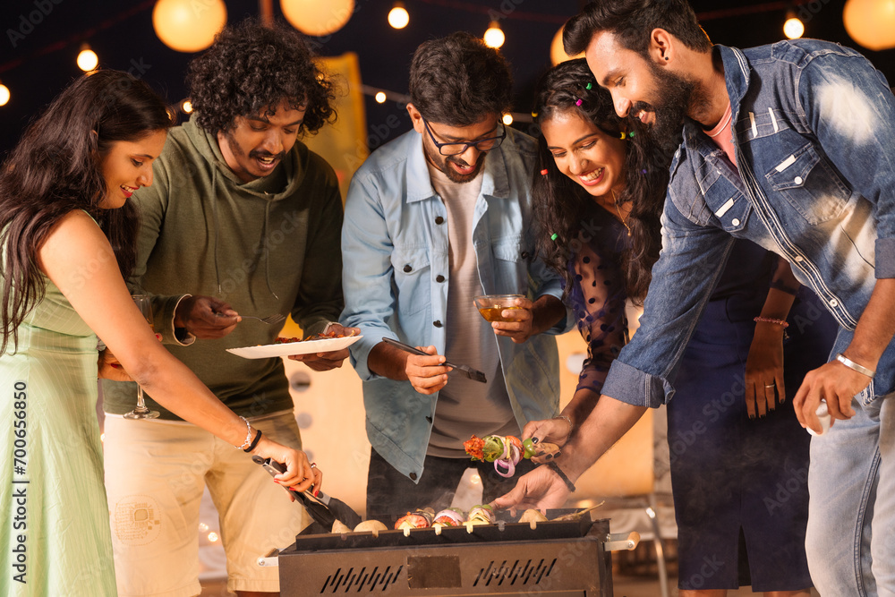 Group of friends busy cooking barbecue grills for dinner during night party - concept of togetherness, birthday gathering and celebration - obrazy, fototapety, plakaty 