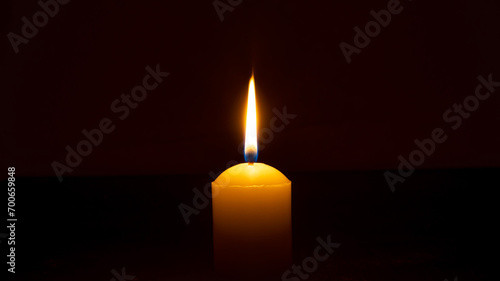 A single burning candle flame or light glowing on a big yellow candle isolated on red or dark background on table in church for Christmas, funeral or memorial service