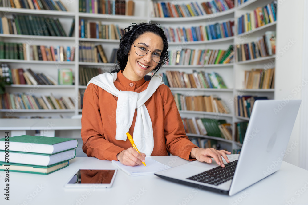 Portrait of a young smiling Latin American female student in a suit studying online at a laptop. He sits at the table in the library, listens and writes the course in a notebook. Portrait of a young smiling Latin American female student in a suit studying online at a laptop. He sits at the table in the library, listens and writes the course in a notebook.