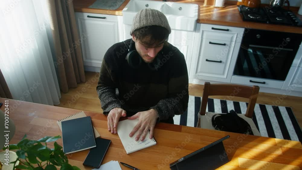 High angle shot of young student man with beanie sitting in modern kitchen tears the page from notebook and throws it back while study with tablet computer and takes notes in notebook High angle shot of young student man with beanie sitting in modern kitchen tears the page from notebook and throws it back while study with tablet computer and takes notes in notebook