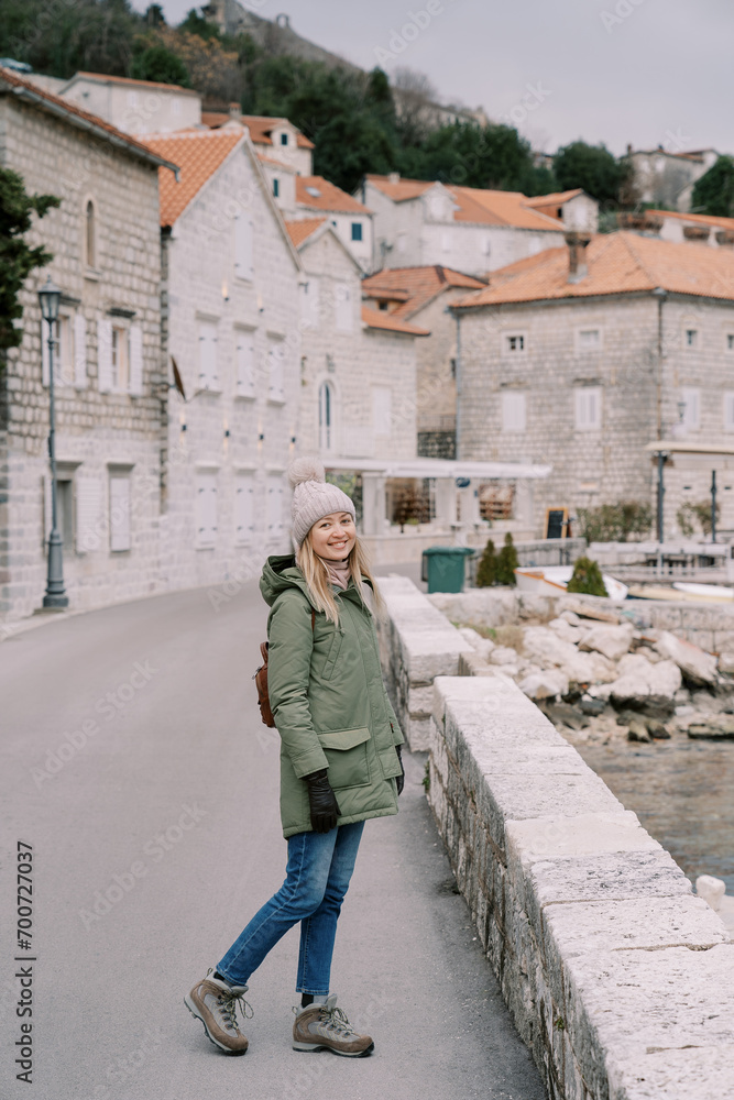 Smiling woman in a jacket with a backpack stands on the Perast embankment. Montenegro