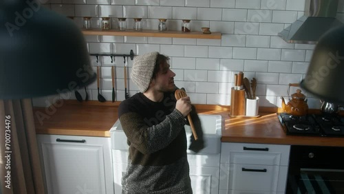 High angle shot of carefree young man with beanie singing into a ladle like a microphone and freely dancing to music in a kitchen at home photo