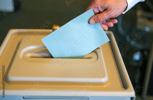 Stimmabgabe im Wahlbüro: Nahaufnahme der Hand eines Mannes, der seinen Stimmzettel in die Wahlurne steckt, viel Copy Space © redaktion93