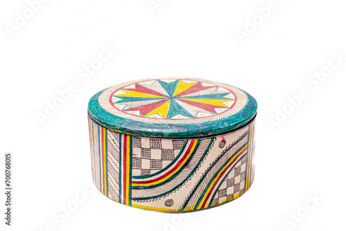 A circular box closed with its lid. With lots of patterns and colors. A checkerboard, curves, stripes, stars and rectangles in beige, black, yellow, red and green. Cut out isolated in white background