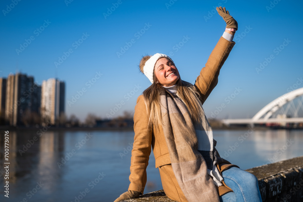 Beautiful woman in warm clothing enjoys resting by the river on a sunny winter day. 