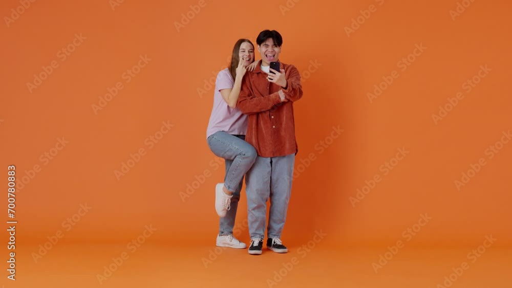 Woman and Asian man take selfie on smartphone by orange wall Woman and Asian man take selfie on smartphone by orange wall