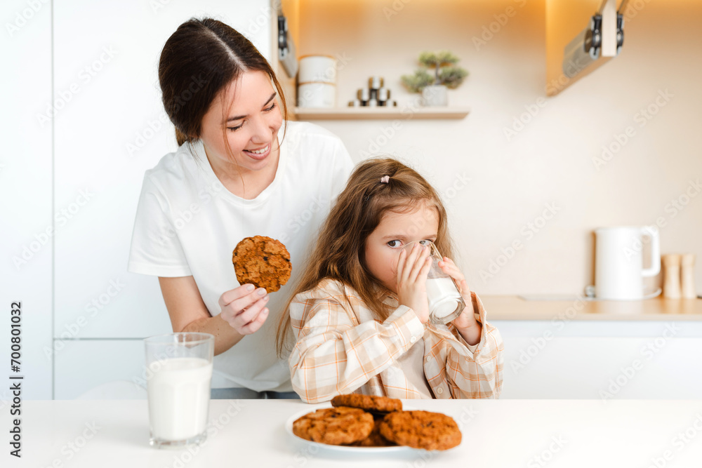 Smiling woman, mother and her little mother holding oatmeal cookies, drinking in milk in kitchen