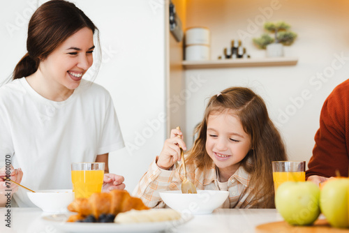 Beautiful young mother and her little daughter having breakfast together sitting at table in kitchen