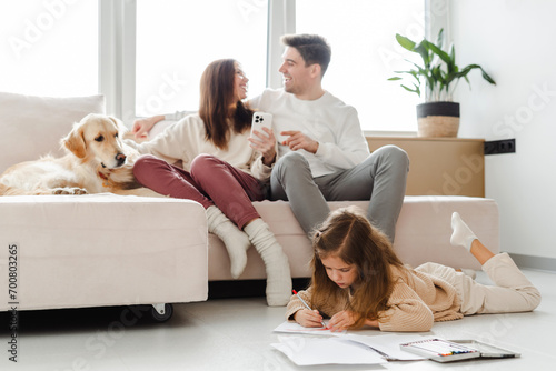 Smiling mother and father, woman holding mobile phone, dog lying on sofa daughter sitting on floor