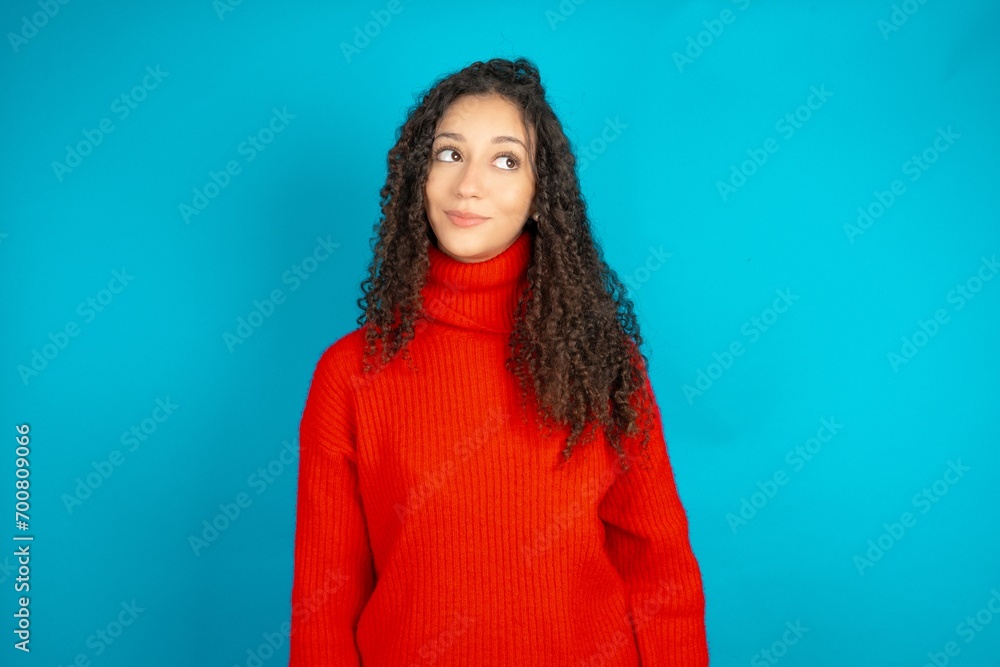 Fototapeta premium Beautiful teen girl wearing knitted red sweater over blue background looking aside into empty space thoughtful