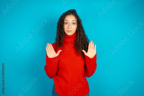 Serious beautiful teen girl wearing red knitted sweater pulls palms towards camera, makes stop gesture, asks to control your emotions and not be nervous photo